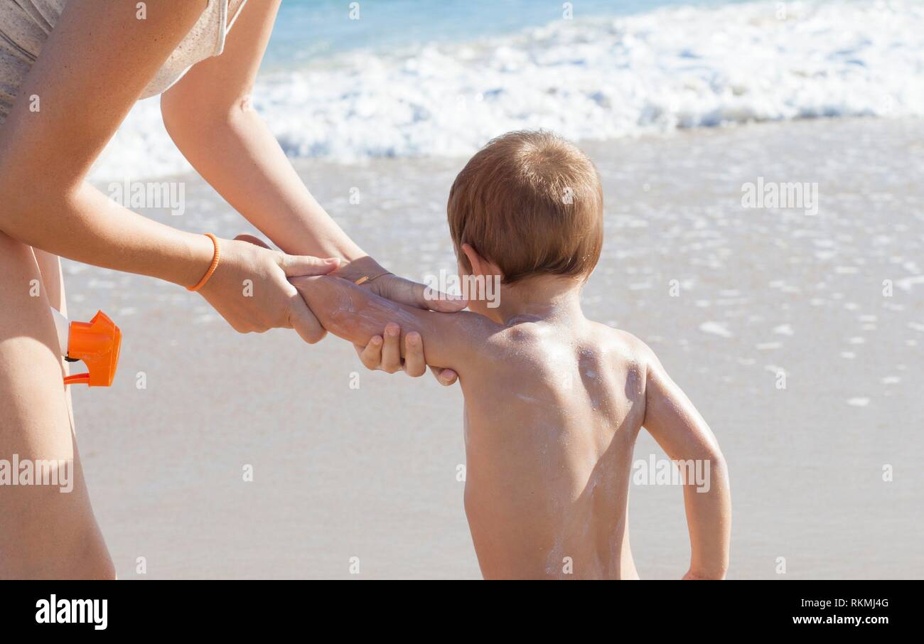Madre l'applicazione di crema di protezione solare per il suo piccolo figlio sul retro. Spiaggia idilliaca sfondo. Foto Stock