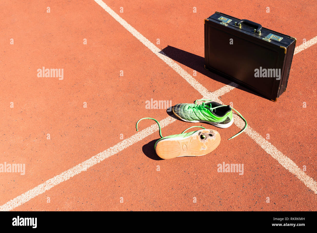 Una ventiquattrore nera e un paio di rotture di green scarpe da corsa su una via di corsa che simbolizza la concorrenza, sfida e burnout nel lavoro e affari. Foto Stock