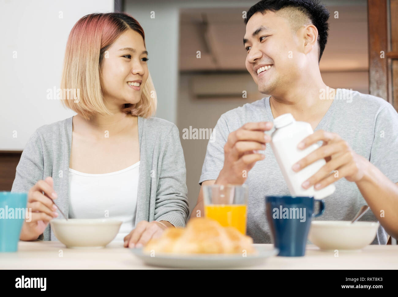 Felice coppia asiatica in pigiama seduta a tavola in cucina a casa la mattina e avente cereale colazione insieme.marito versare il latte nella tazza moglie Foto Stock
