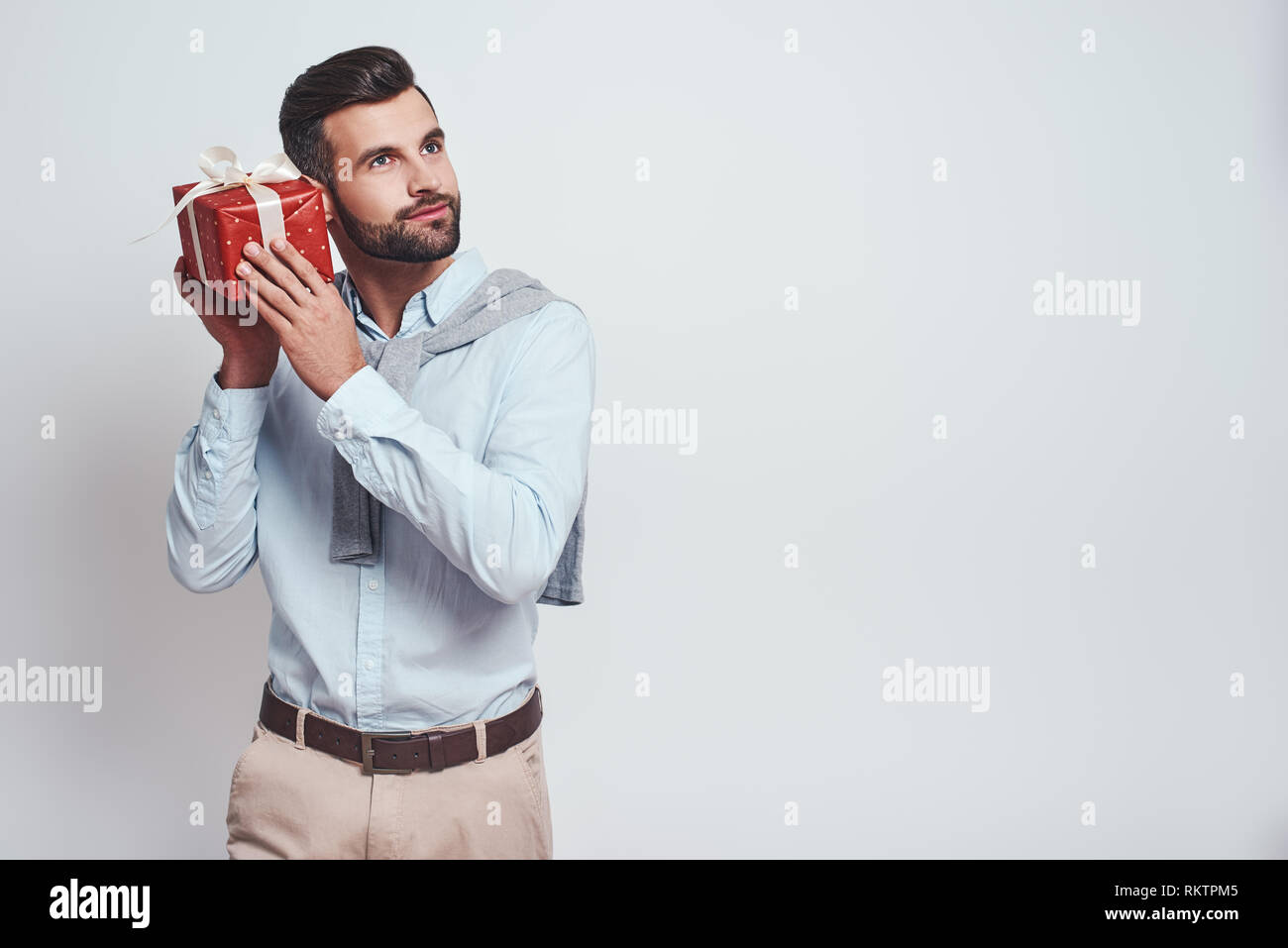 Che cos'è all'interno? Attraente uomo elegante è in possesso di una confezione regalo vicino al suo orecchio e cercando di indovinare che cosa è interno mentre in piedi su sfondo grigio. Close-up immagine Foto Stock