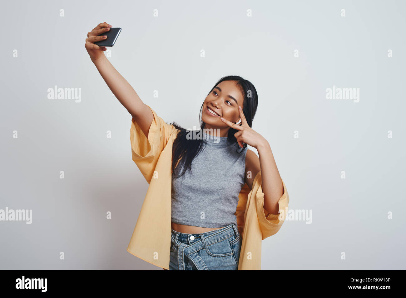 Tempo Selfie bella giovane donna asiatica in abbigliamento casual sorridente e gesticolando mentre si effettua una selfie in studio su sfondo grigio. Close up ritratto. Concetto di tecnologia Foto Stock