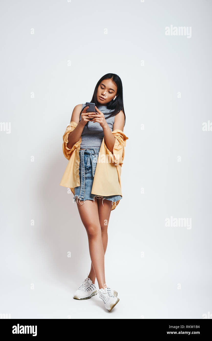 Navigare in rete. A piena lunghezza Ritratto di bella donna asiatica in abbigliamento casual guardando il suo smart phone mentre in piedi su sfondo grigio. Concetto di tecnologia Foto Stock