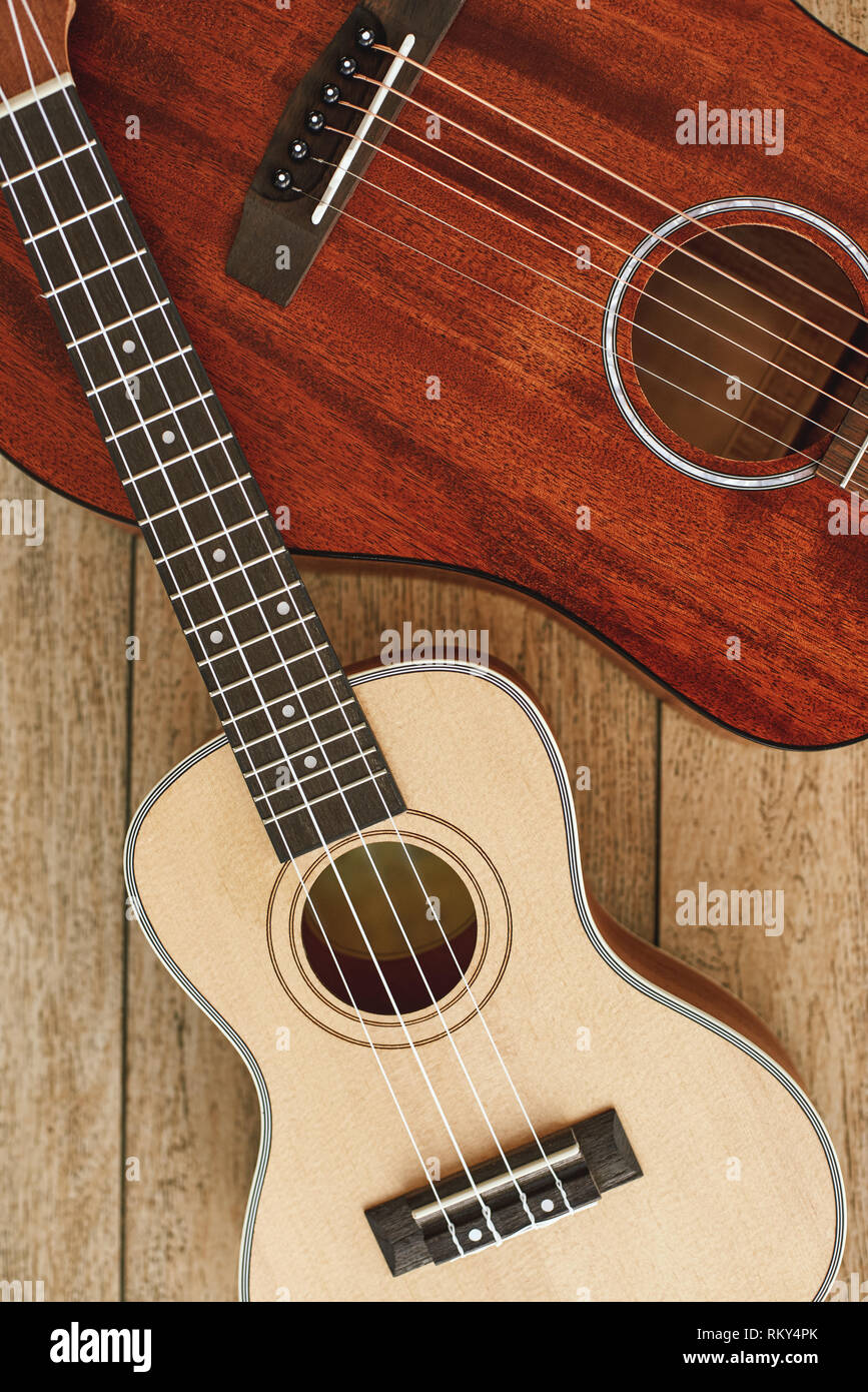 Tema per chitarra. Verticale in alto vista di acustica e ukulele chitarre giacente su ogni altra contro il pavimento in legno. Il concetto di musica. Strumenti musicali Foto Stock