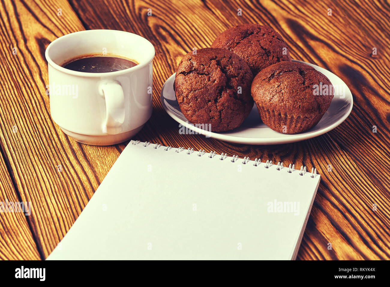 Diversi muffin al cioccolato con pasticceria scuro con una tazza di caffè e un blocco note su un tavolo di legno. Foto Stock