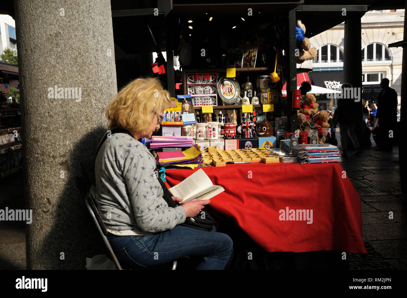Un senior donna trascorrere un momento tranquillo la lettura di un libro a lei di stallo del mercato turistico di memorabilia, Covent Garden, Londra, Inghilterra, Regno Unito Foto Stock