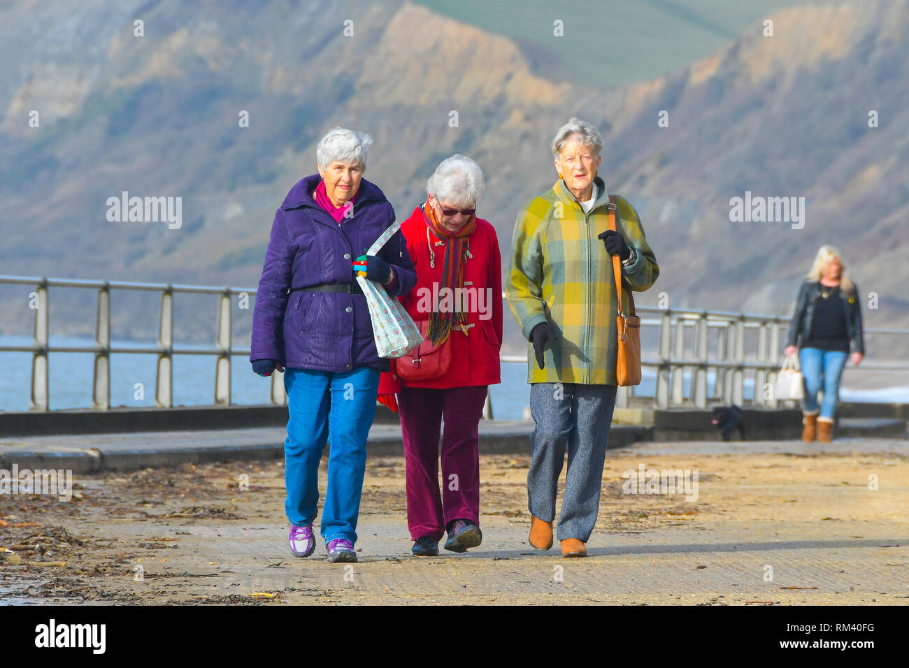 West Bay, Dorset, Regno Unito. Il 13 febbraio 2019. Regno Unito Meteo. Fumoso sole riscalda tre onorevoli colleghe che stanno camminando lungo il lungomare di West Bay nel Dorset che sono avvolti ben dopo una fredda per iniziare la giornata. Le temperature sono impostate a salire come venti da sud dai tropici sono impostati a soffiare di tutta la contea nel giro di un paio di giorni portando temperature sopra la media. Credito Foto: Graham Hunt/Alamy Live News Foto Stock