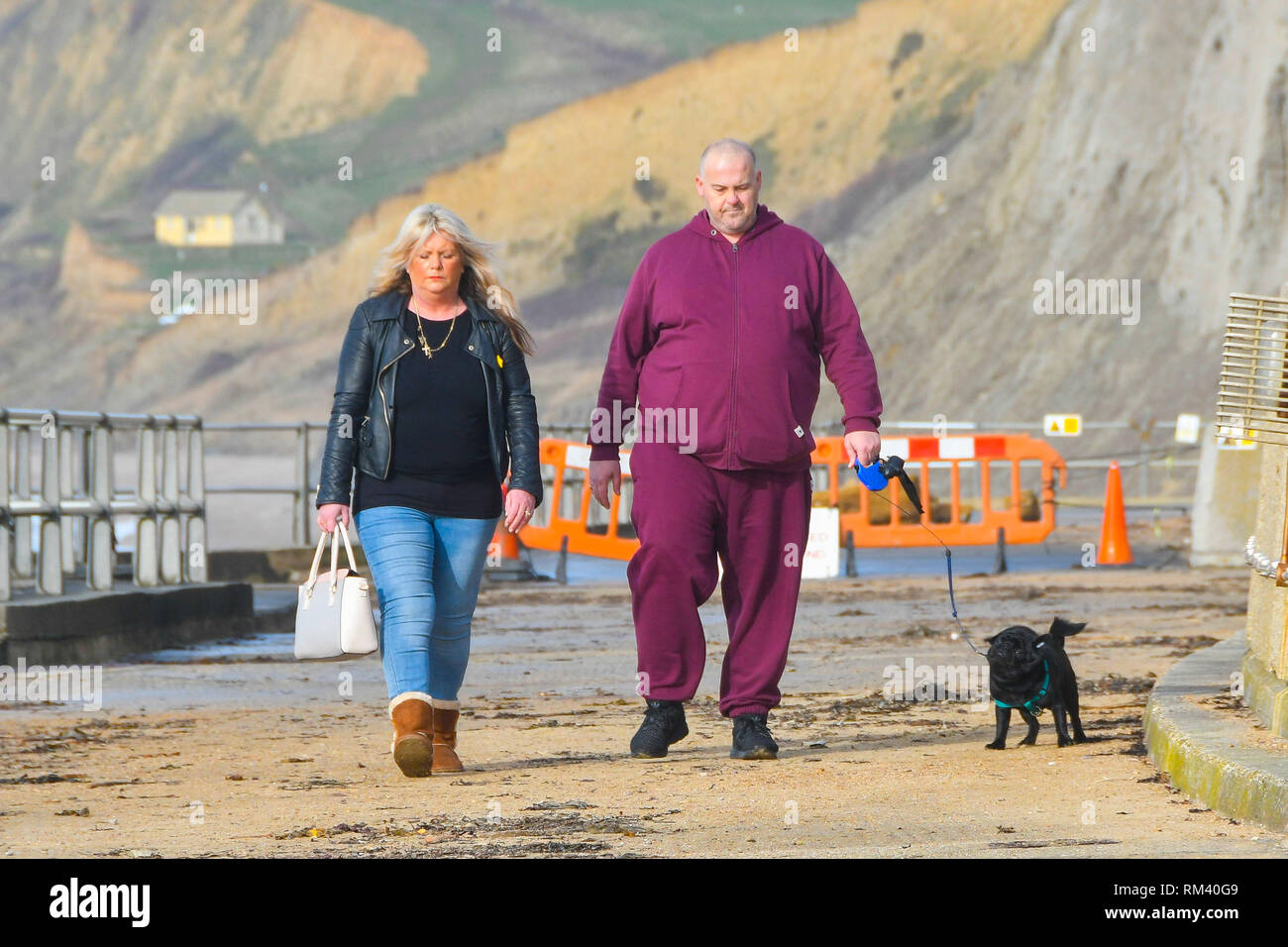 West Bay, Dorset, Regno Unito. Il 13 febbraio 2019. Regno Unito Meteo. Fumoso sole riscalda cane escursionisti che stanno camminando lungo il lungomare di West Bay nel Dorset dopo una fredda per iniziare la giornata. Le temperature sono impostate a salire come venti da sud dai tropici sono impostati a soffiare di tutta la contea nel giro di un paio di giorni portando temperature sopra la media. Credito Foto: Graham Hunt/Alamy Live News Foto Stock