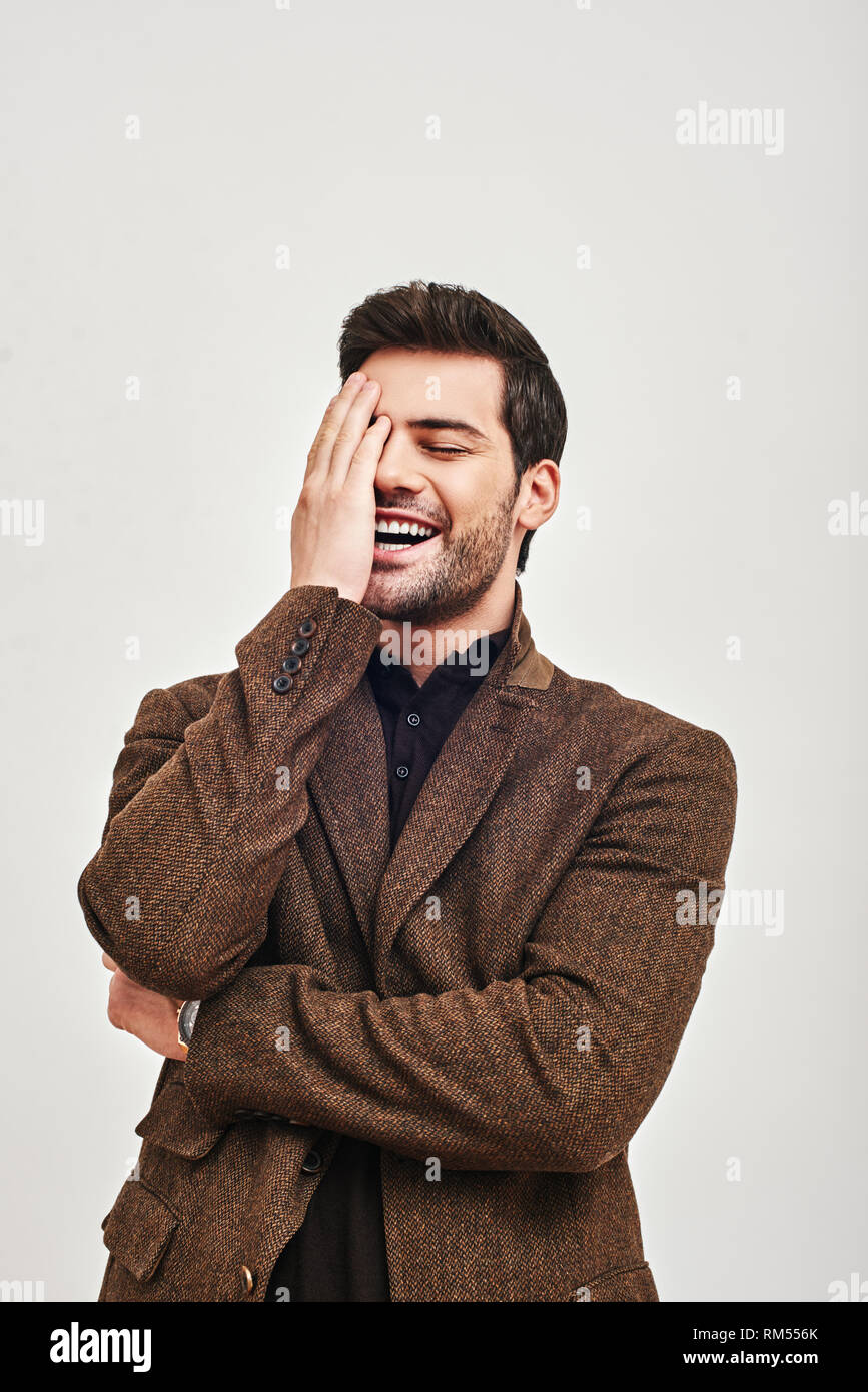 Ritratto di un giovane uomo bello indossare giacca marrone, che copre il volto con le mani e ridere isolato su uno sfondo bianco. Concetto di felicità Foto Stock