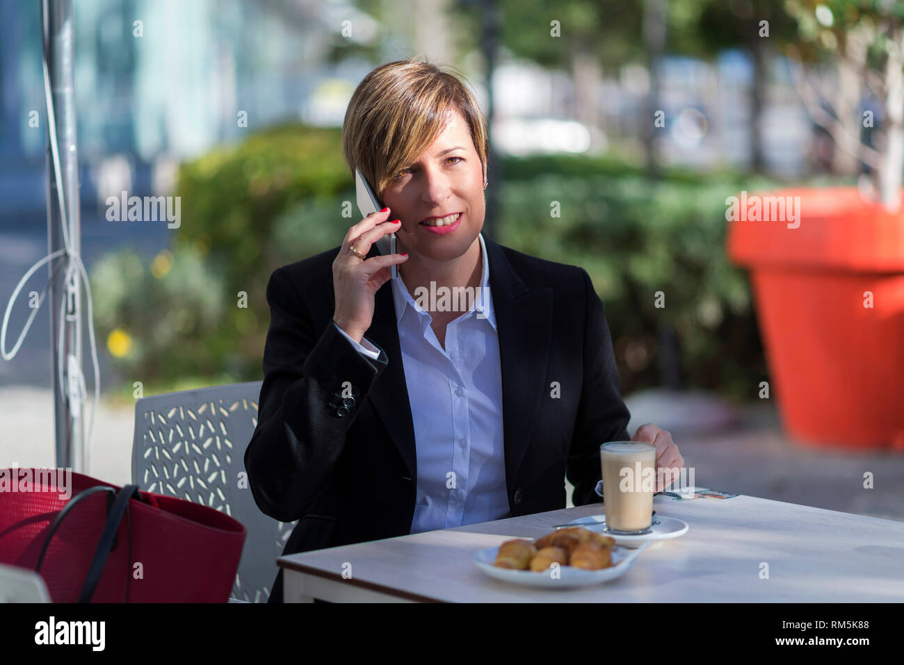 Vista frontale di un elegante business sorridente donna seduta su una sedia in un caffè mentre utilizzando un telefono cellulare godendo la conversazione all'aperto Foto Stock