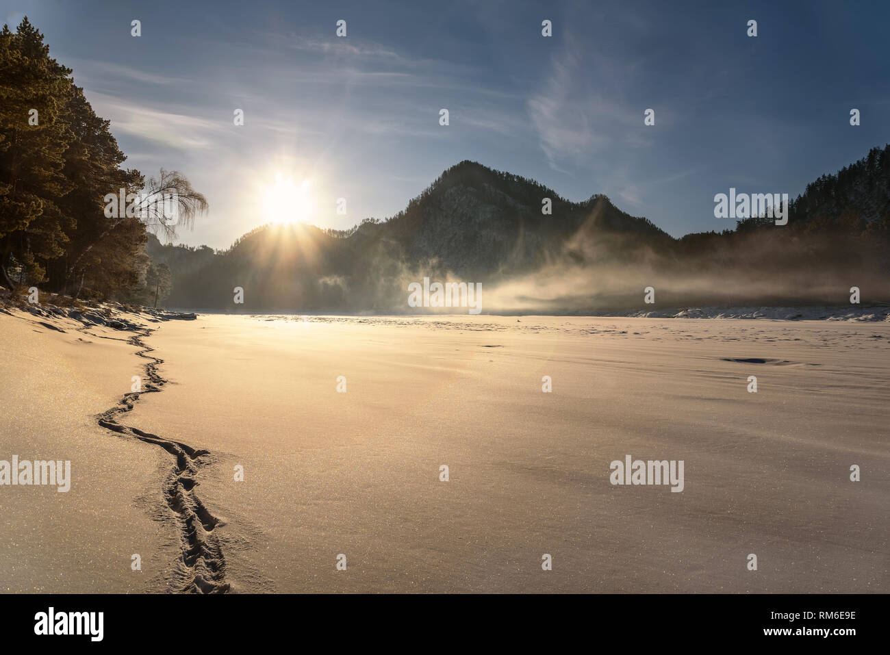 Incantevole paesaggio invernale con montagne, sun, fiume congelato sotto la neve, nebbia e Impronte sulla neve lungo la riva Foto Stock