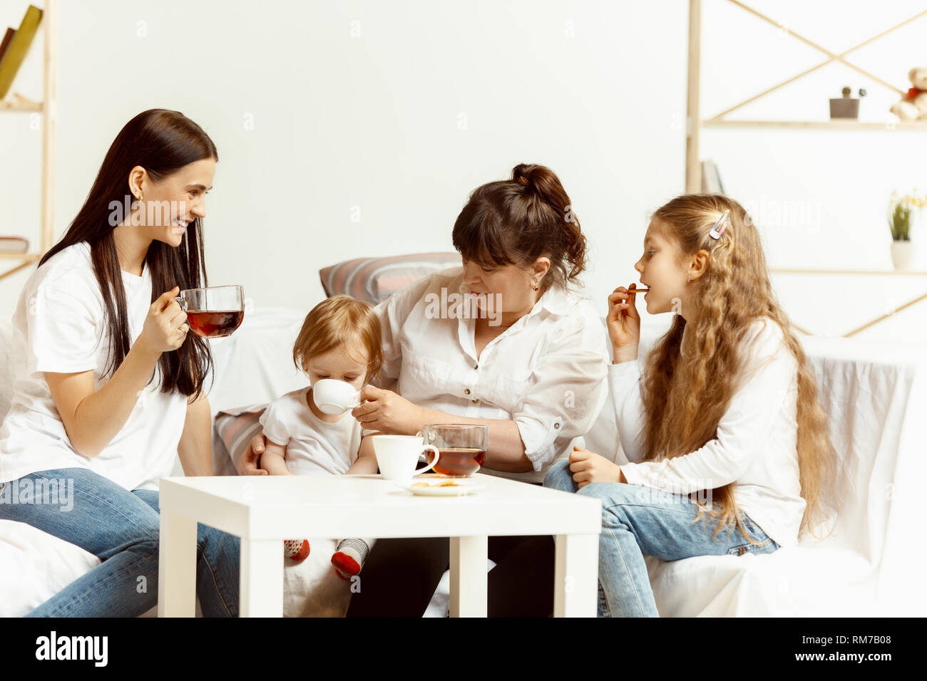 Due bambine loro attraente giovane madre e il loro affascinante nonna seduti sul divano e trascorrere del tempo insieme a casa. Generazione di donne. La Giornata internazionale della donna. Felice Festa della mamma. Foto Stock