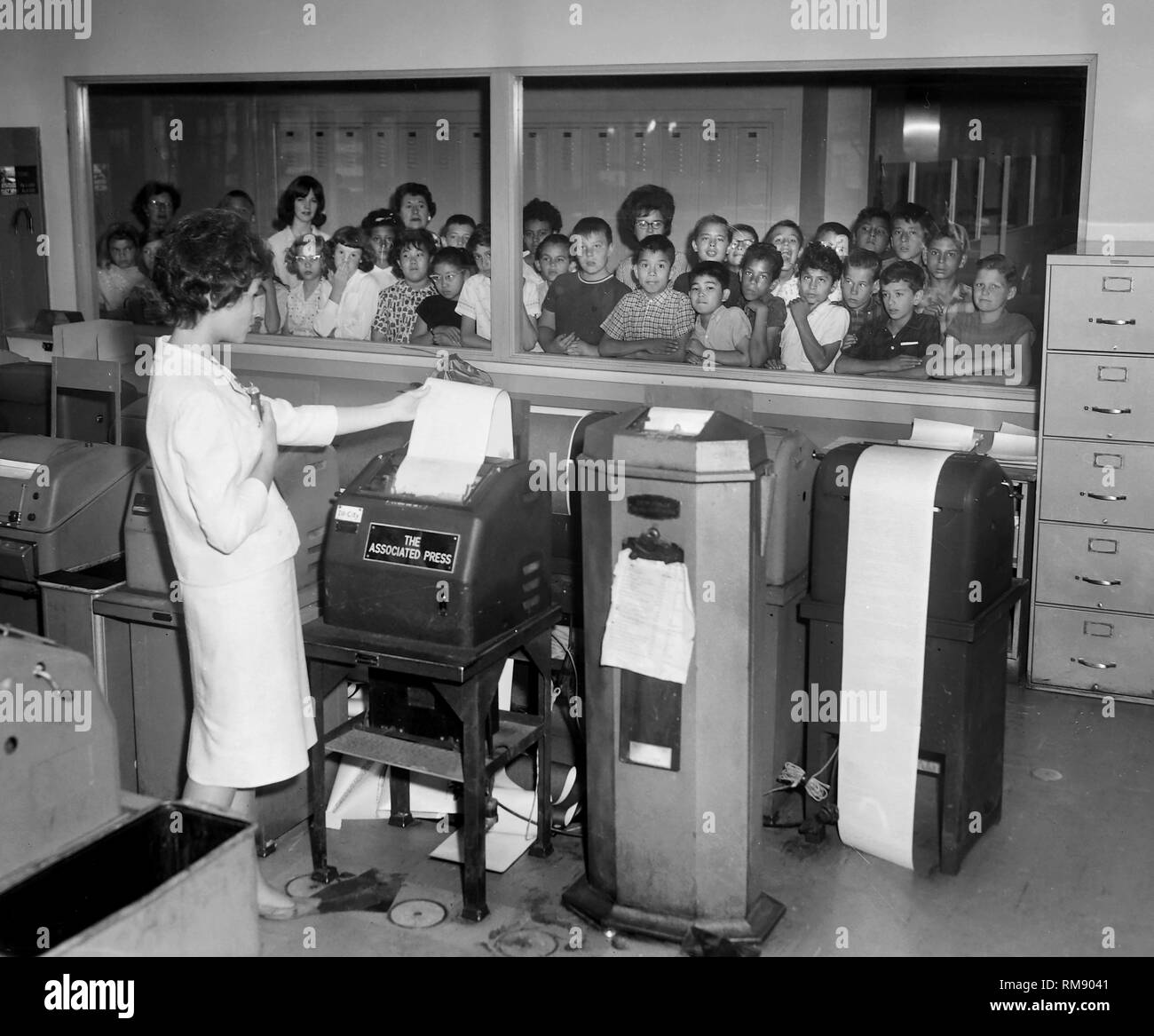Una touring gruppo di bambini in età scolare su una gita di guardare attraverso una finestra come una donna raccoglie copia della trasmissione dal filo macchine per la modifica di una newsroom di Chicago, ca. 1965. Foto Stock