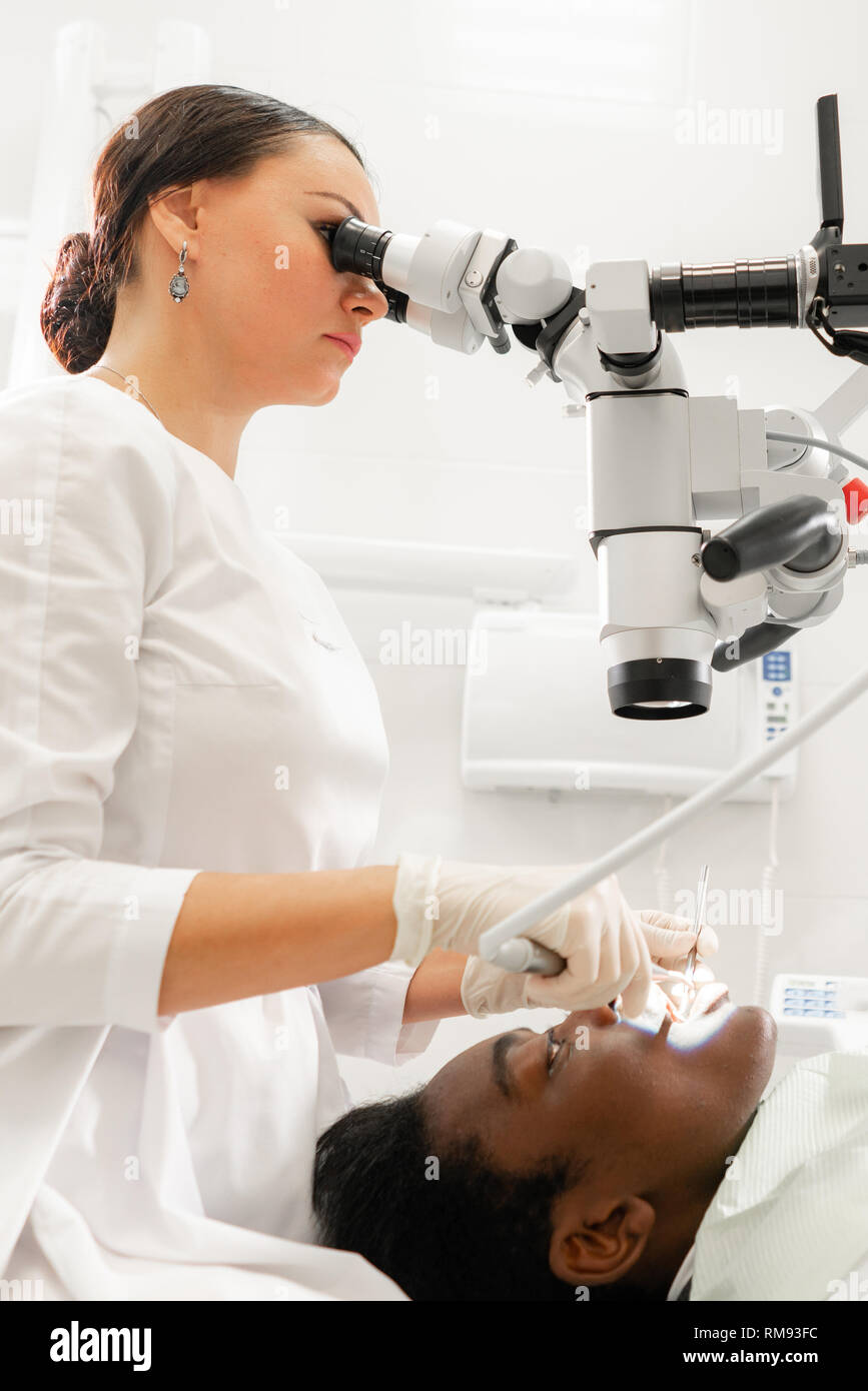 Giovane donna dentista il trattamento di canali radicolari mediante microscopio nella clinica dentale. Uomo paziente sdraiato sulla poltrona del dentista con la bocca aperta. Medicina Foto Stock