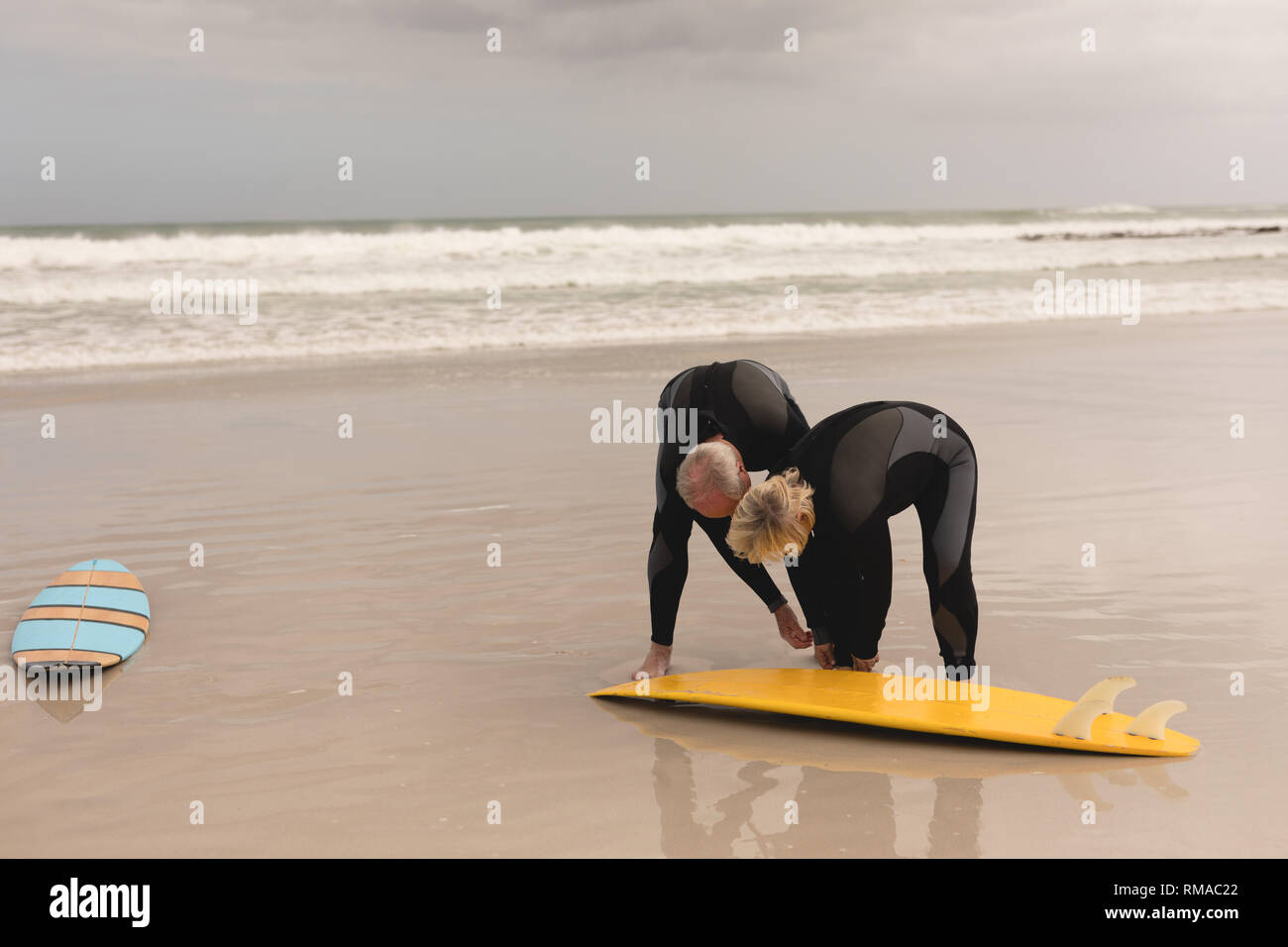 Senior aiuta l uomo donna senior di indossare le tavole da surf guinzaglio sulla spiaggia Foto Stock