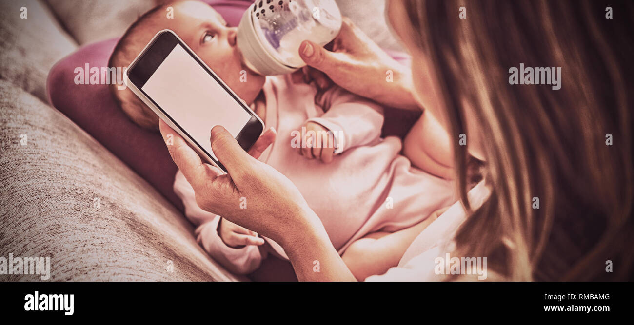 Madre utilizzando il telefono cellulare mentre si alimenta il suo bambino con bottiglia di latte Foto Stock