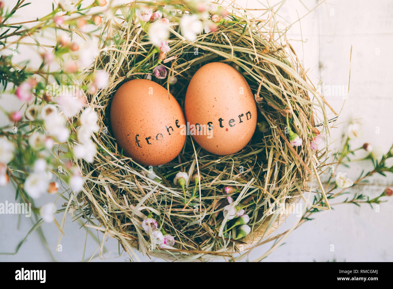 Le uova di pasqua con il timbro di gomma 'Frohe Ostern' Testo messaggio di saluto in un fieno e fiori nido su un bianco tavolo in legno Foto Stock