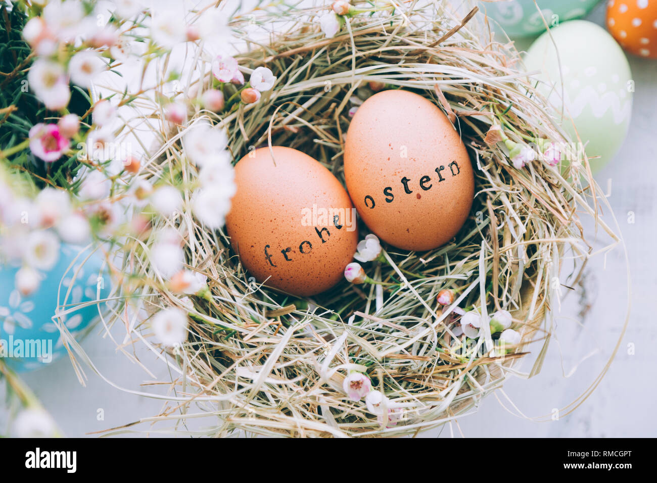 Le uova di pasqua con il timbro di gomma 'Frohe Ostern' Testo messaggio di saluto in un fieno e fiori su un bianco sullo sfondo di legno Foto Stock
