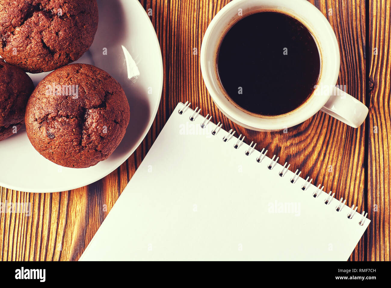 Diversi muffin al cioccolato con pasticceria scuro con una tazza di caffè e un blocco note su un tavolo di legno. Foto Stock