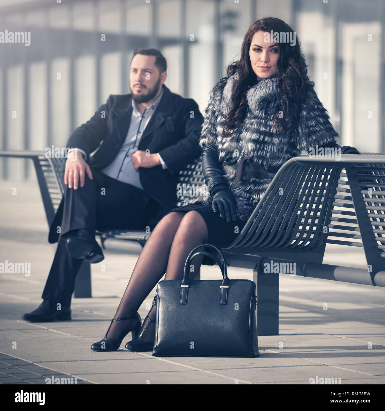 Moda giovane coppia in conflitto seduta sul banco di prova su strada di città elegante alla moda uomo nel classico mantello nero e la donna che indossa la pelliccia di volpe camicia Foto Stock