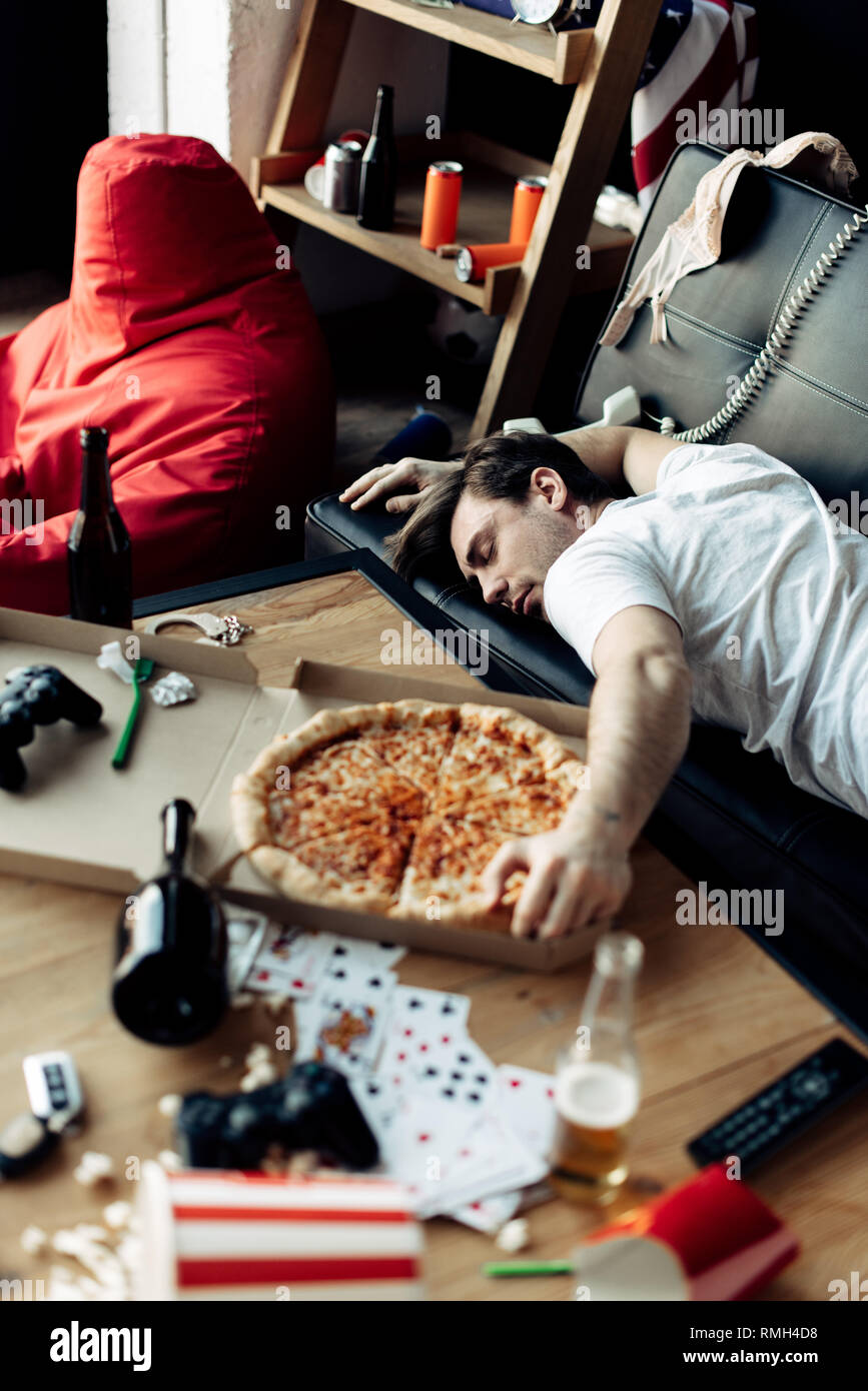 Uomo ubriaco che dorme sul divano vicino a una gustosa pizza al disordine home after party Foto Stock