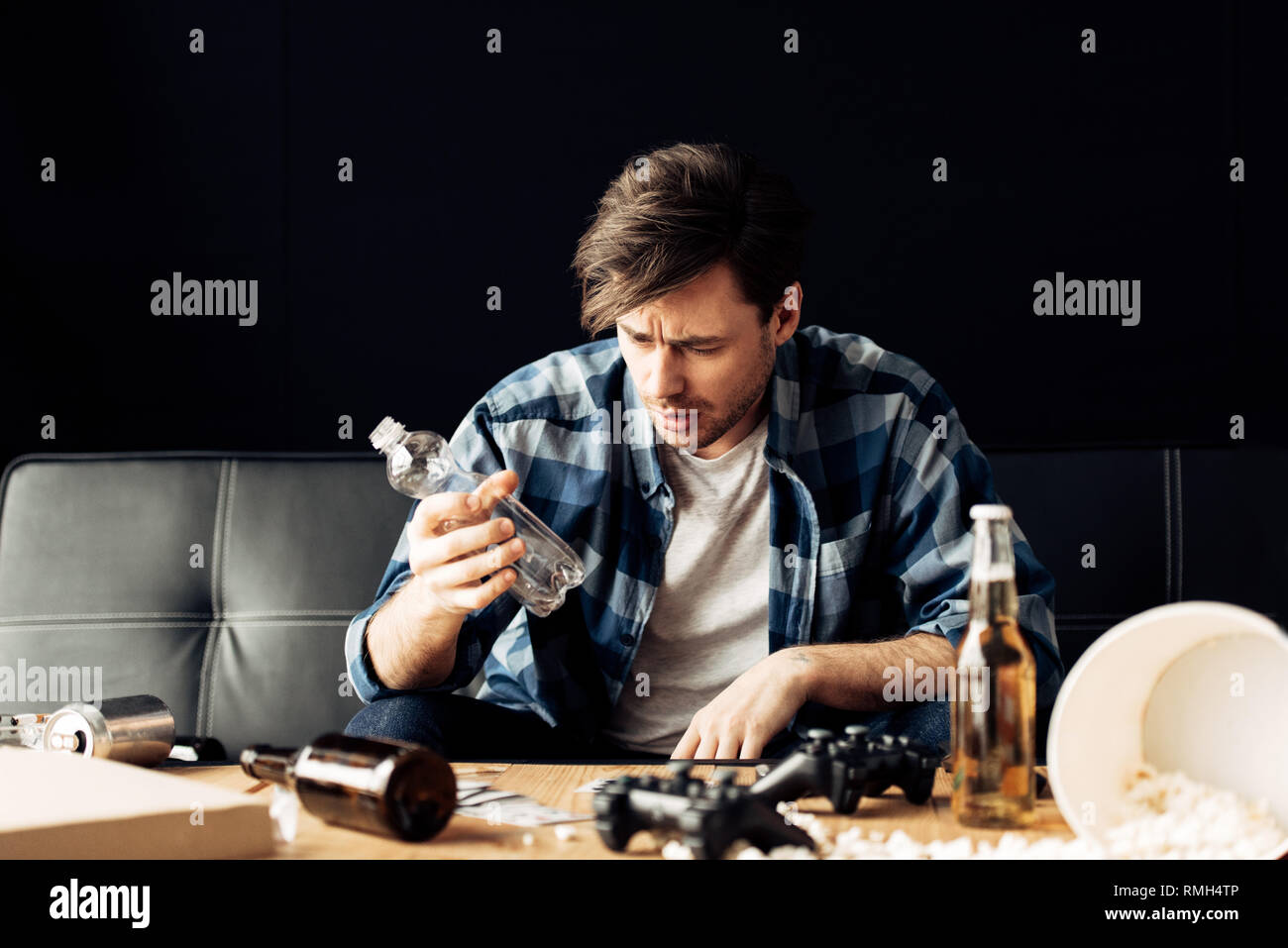 Bel uomo che soffre di sbornia guardando bottiglia vuota mentre è seduto sul divano Foto Stock