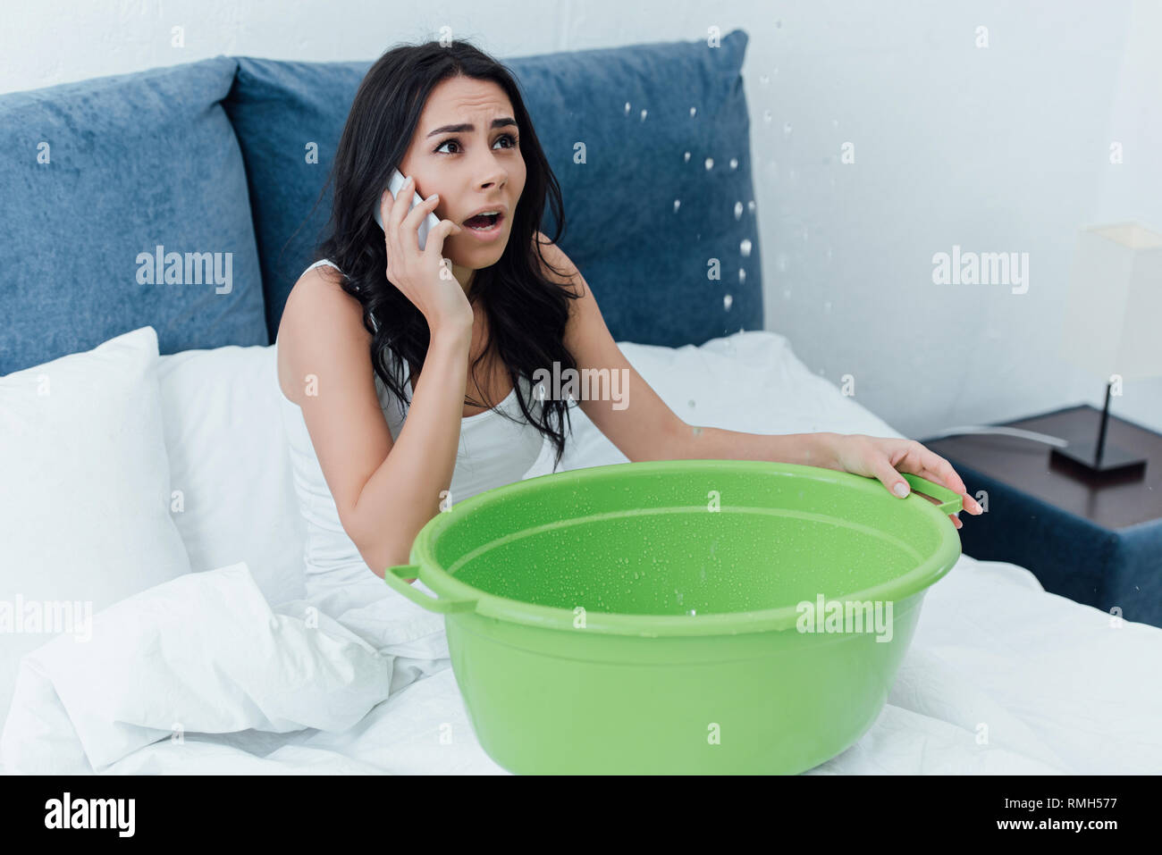 Frightened donna parlando su smartphone e guardando la perdita di acqua in camera da letto Foto Stock