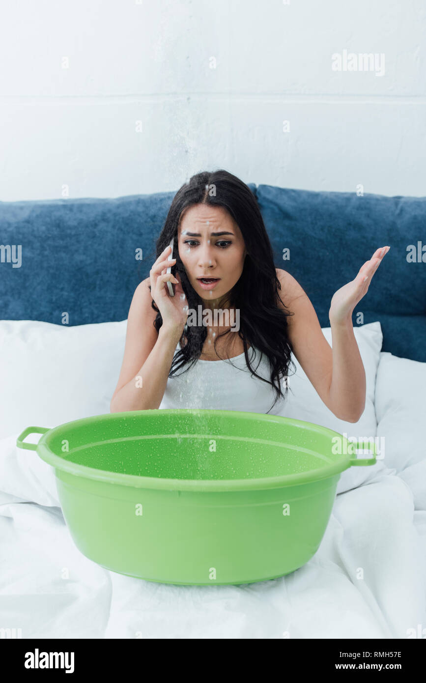 Scioccato donna parlando sullo smartphone mentre si ha a che fare con la perdita di acqua in camera da letto Foto Stock