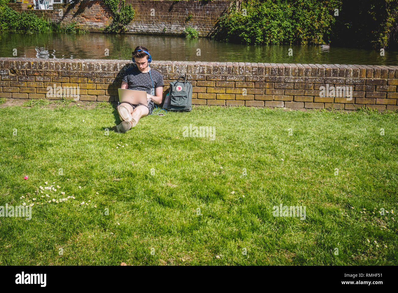 Canterbury, Regno Unito - Maggio, 2018. Un giovane uomo che lavora su un computer portatile e di ascoltare musica mentre è seduto sul prato di un parco. Foto Stock