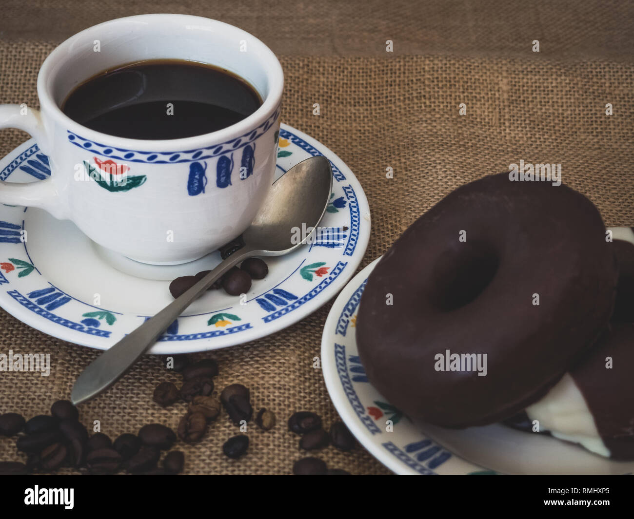 La prima colazione di una tazza in porcellana, un antico cucchiaio, ciambelle al cioccolato e caffè in grani su una tela vintage background Foto Stock
