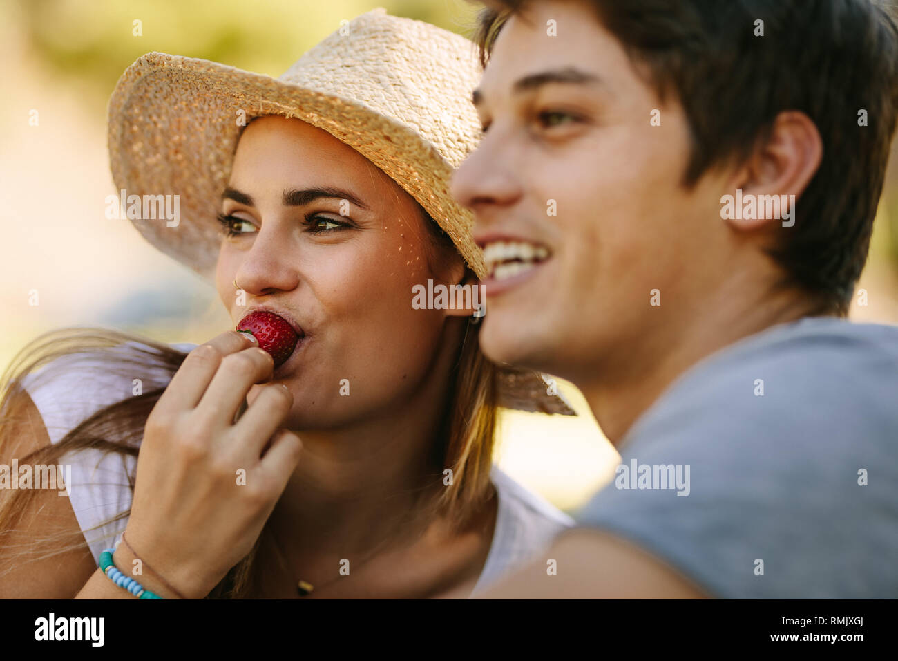 Coppia sorridente seduti insieme all'aperto e guardando lontano. Donna in hat mangiare una fragola seduto con il suo ragazzo in un parco. Foto Stock