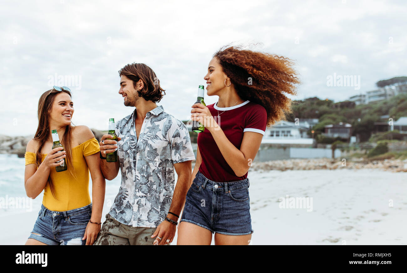 Gruppo di tre giovani amici passeggiate all'aperto con bottiglie di birra. Multi-etnico amici con birre camminando lungo la spiaggia. Foto Stock