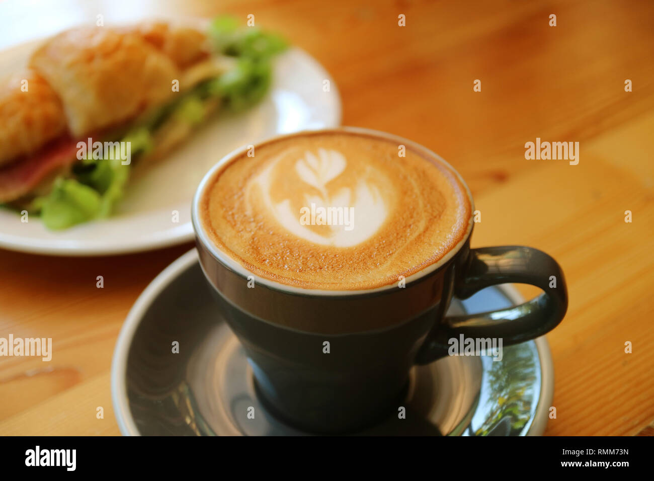 Una tazza di cappuccino caldo il caffè in un grigio scuro tazza servita su un tavolo di legno con croissant sfocate Sandwich in background Foto Stock