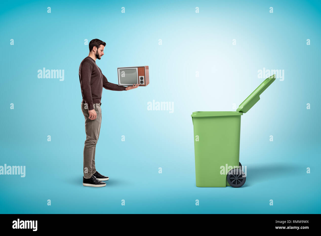 Vista laterale di un uomo in un abbigliamento casual in piedi accanto a un cestino aperto possibile e tenendo un po' vecchio televisore. Foto Stock