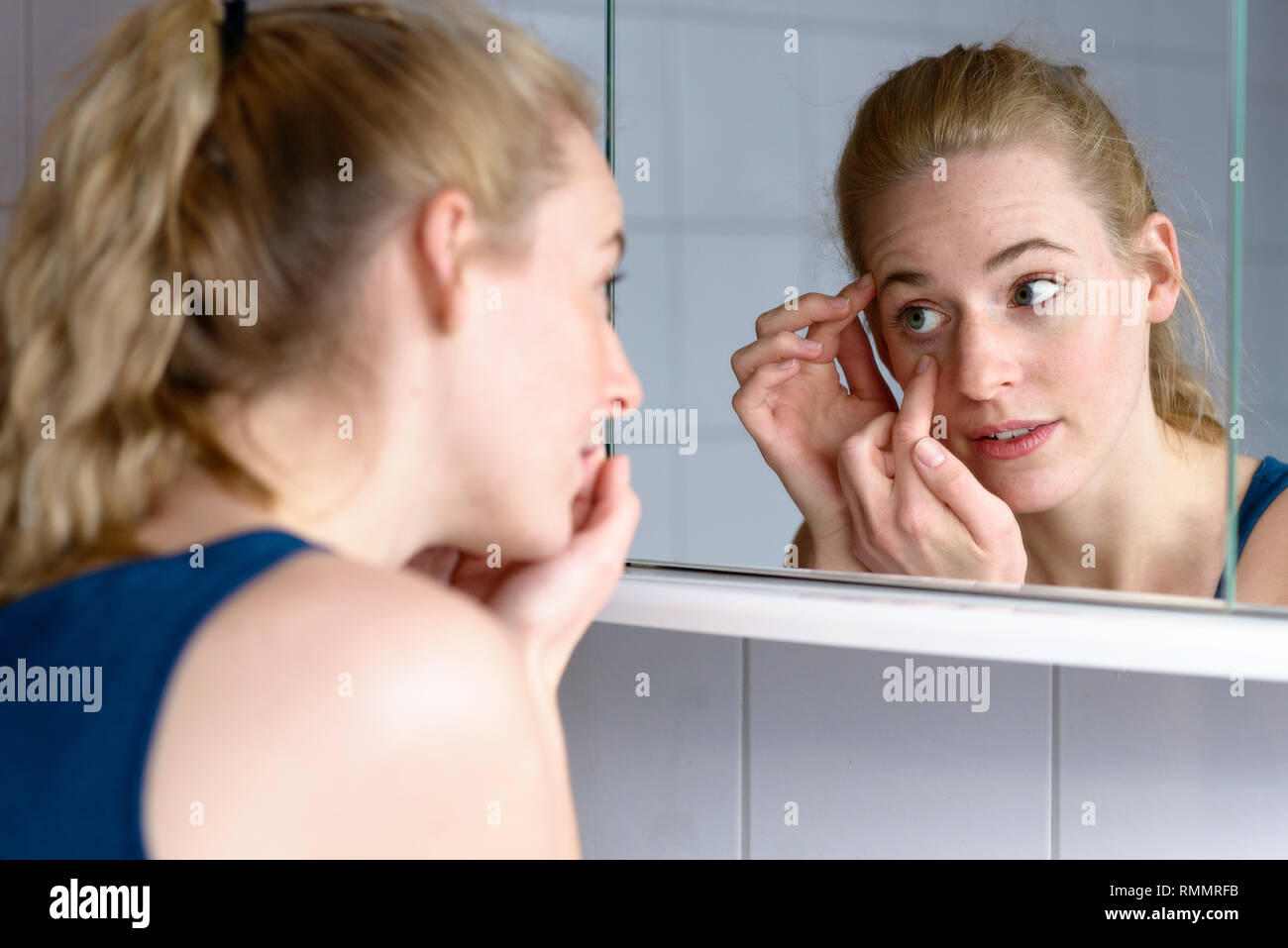 Al di sopra della spalla di una giovane e bella donna preoccupati di invecchiamento mentre guardando nello specchio a la pelle intorno agli occhi Foto Stock