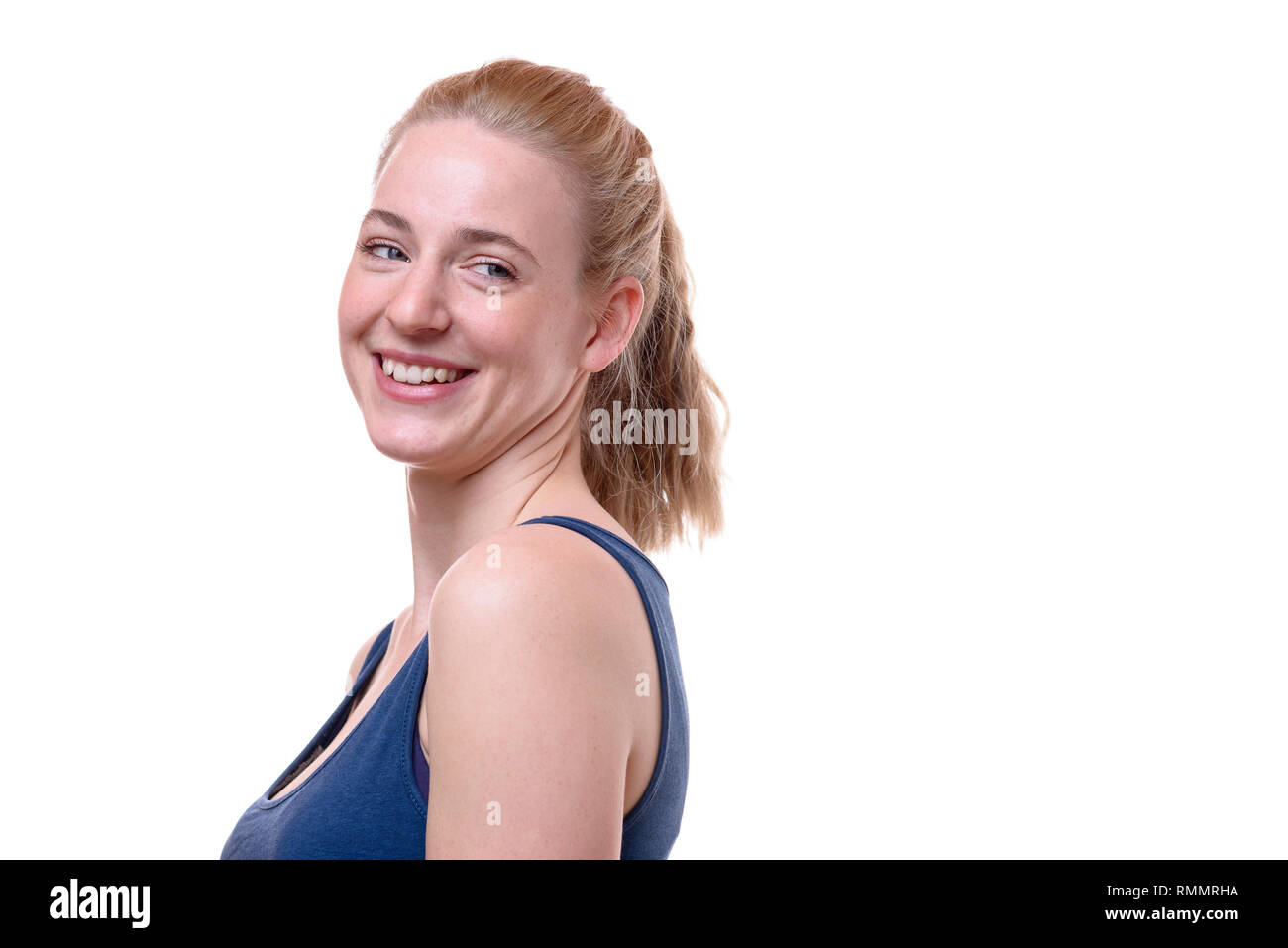 Felice vivace giovane donna bionda volgendo lo sguardo sopra la sua spalla con un sorriso raggiante isolato su bianco con spazio di copia Foto Stock