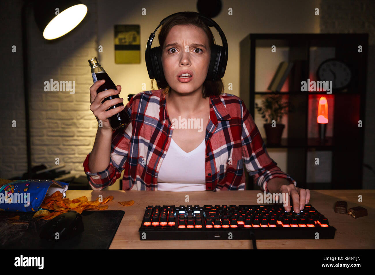 Ragazza eccitato che indossa la cuffia durante il gioco online su computer, mangiando snack Foto Stock