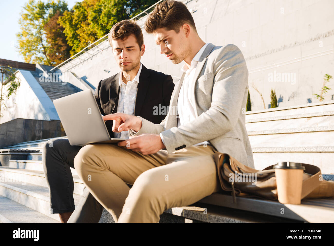 Foto di maturo uomini d'affari in giacca seduti sulla scalinata esterna e utilizzando computer portatile durante la giornata di sole Foto Stock