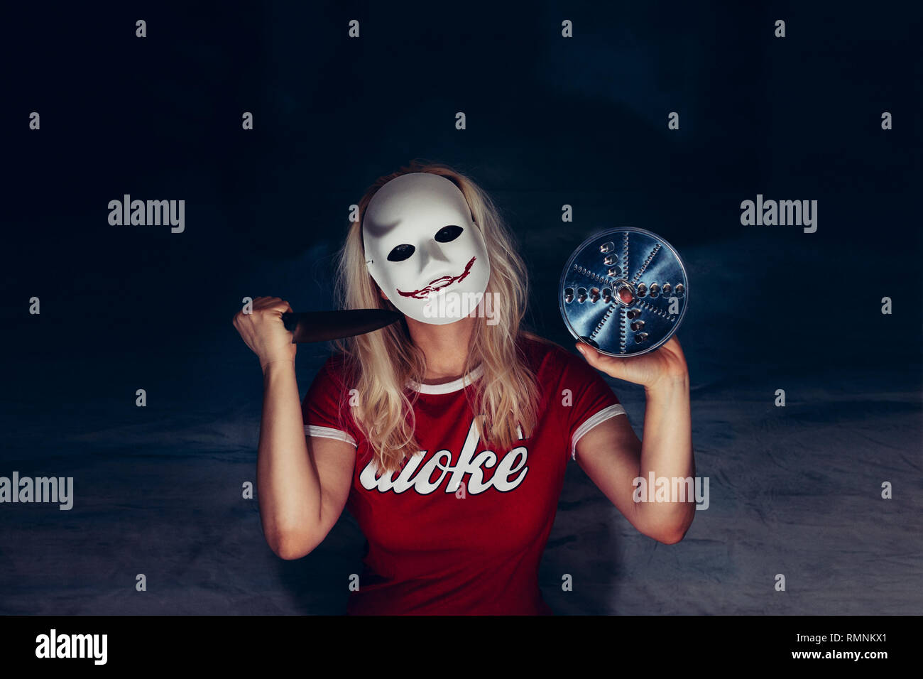 Donna bionda sotto la maschera bianca con spooky sanguinosa sorriso, tenendo il coltello e la pala in difesa di auto, concetto di orrore, indossa una t-shirt rossa con lettera del bianco Foto Stock