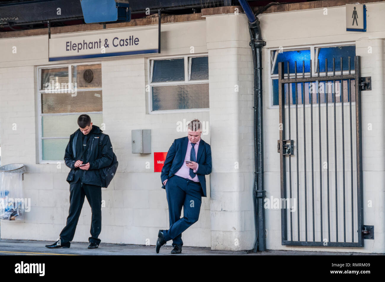 Due passeggeri guardando i loro telefoni cellulari sulla piattaforma a Elephant & Castle dalla stazione di Londra Sud, in attesa di un treno. Foto Stock