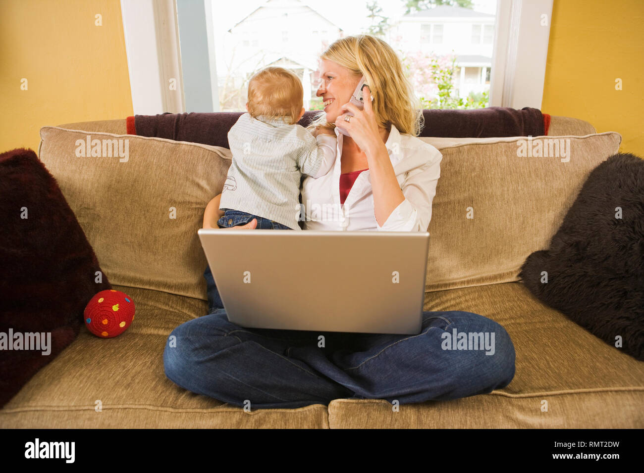 Un mnid-30s Caucasian madre detiene il suo curioso figlio mentre si parla al telefono davanti a un computer portatile. Foto Stock