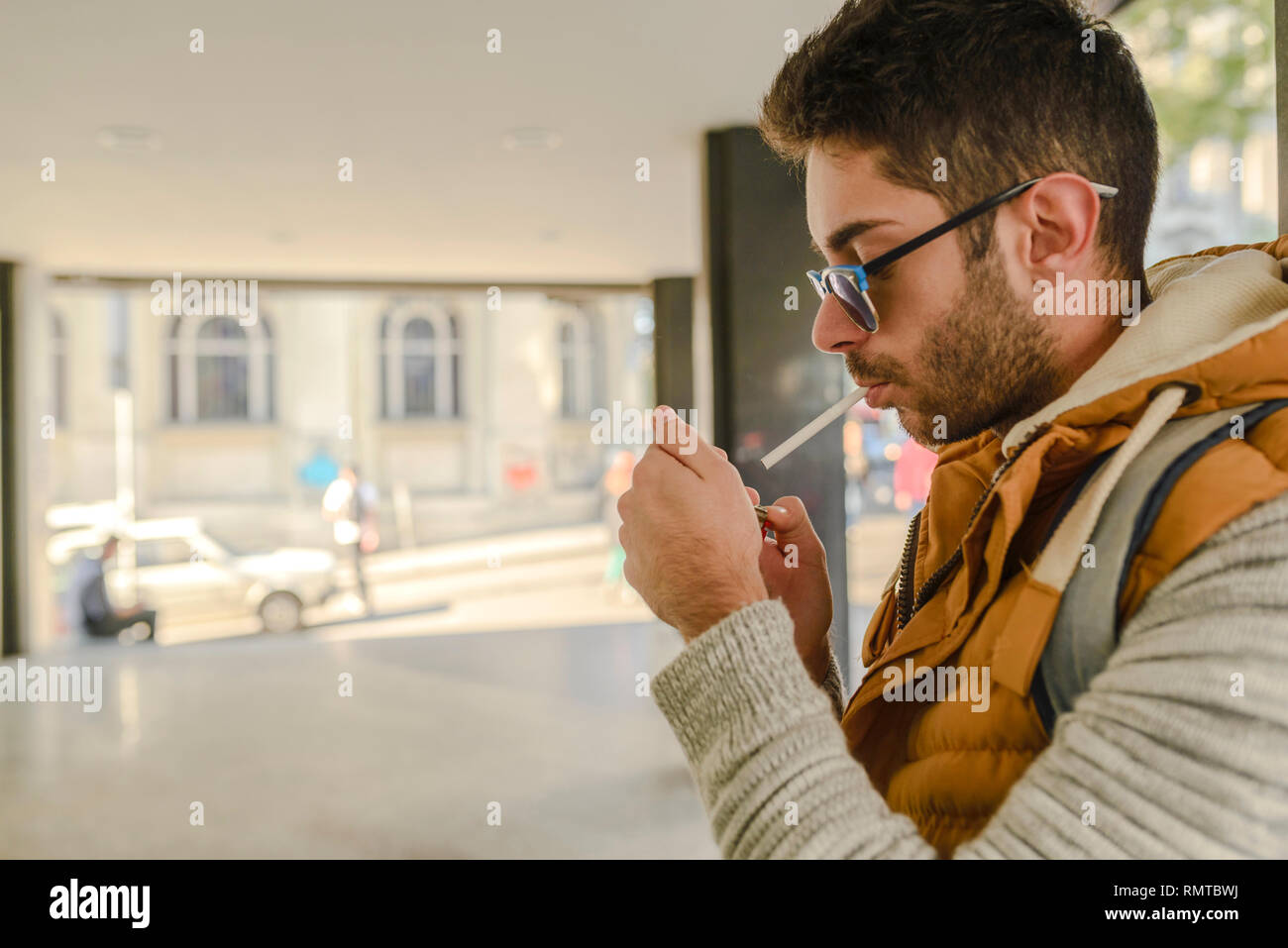 Giovane bello hipster con camicia arancione e occhiali da sole godendo di una sigaretta in strada. Foto Stock