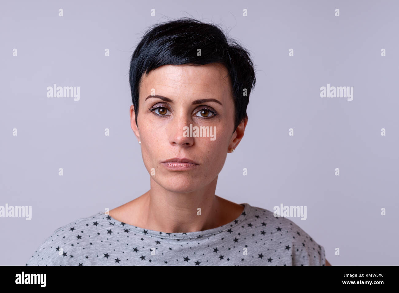 Attraente donna alla moda con corti capelli scuri guardando attentamente la fotocamera su grigio Foto Stock