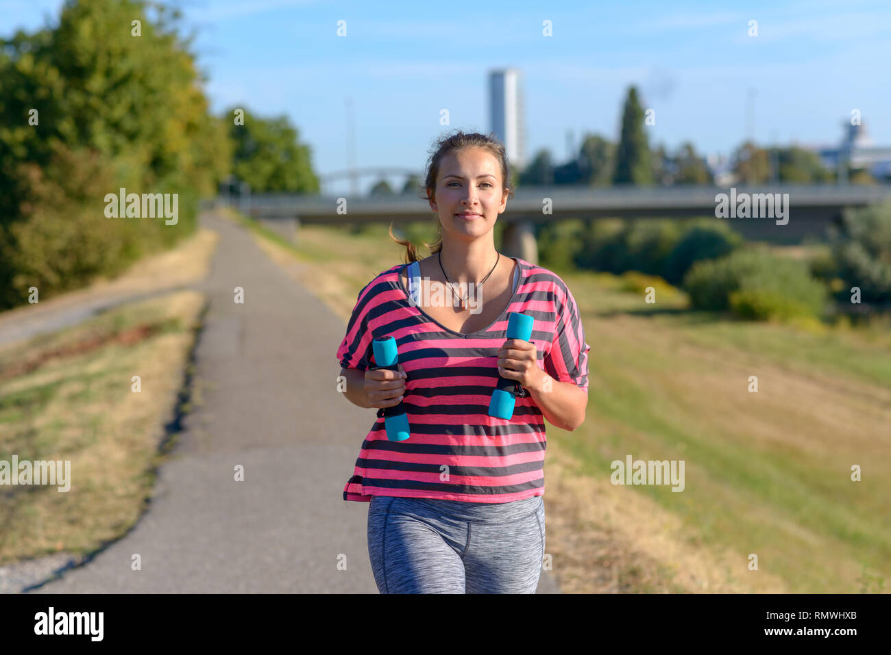 Montare la giovane donna jogging tenendo piccoli pesi a manubrio nelle sue mani lungo una strada rurale sorridente felicemente come lei si avvicina alla telecamera mentre godendo della sua Foto Stock