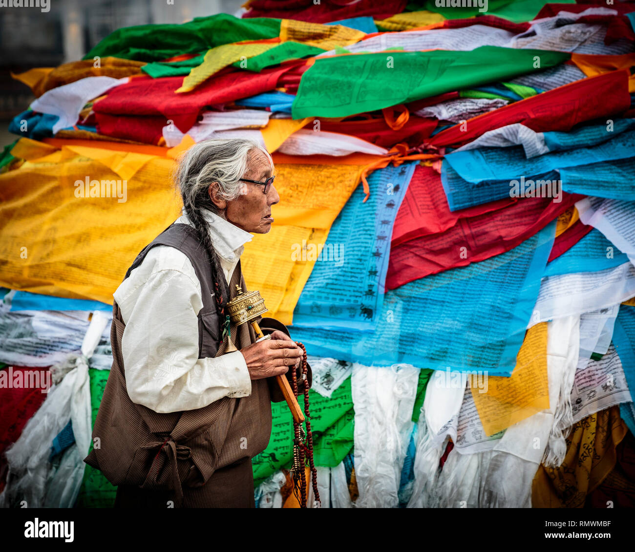 Lhasa, in Tibet. Il 20 luglio 2016. Donna tibetana spining il suo palmare ruota di preghiera passeggiate passato un solido background di colorate bandiere di preghiera. Foto Stock