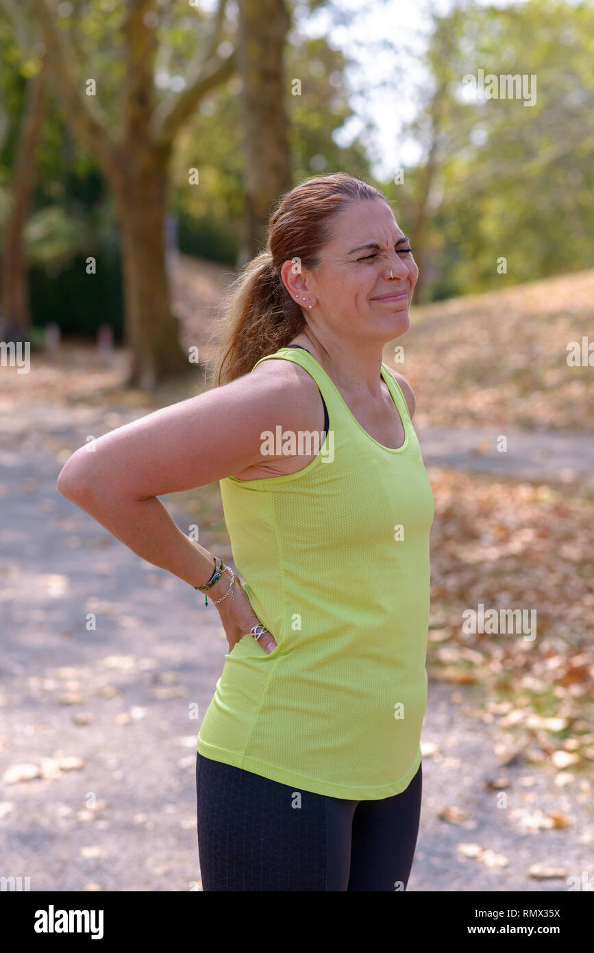 Donna di mezza età jogging subisce un infortunio muscolare in piedi tenendo la sua parte posteriore inferiore mentre smorfie nel dolore, vicino la parte superiore del corpo vista Foto Stock