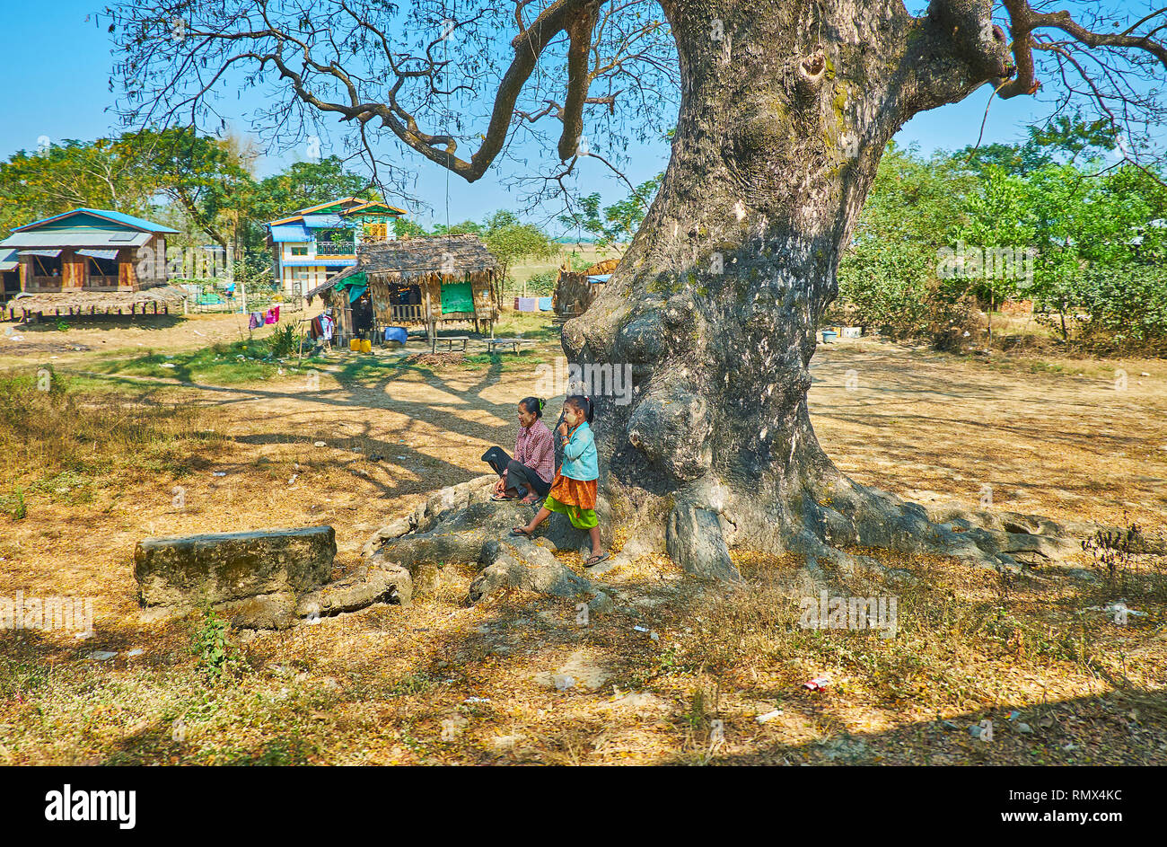 DAR PEIN, MYANMAR - 15 febbraio 2018: gli abitanti del villaggio sedersi sotto il vecchio albero dalla ferrovia con una vista sul stilt capanne sullo sfondo, su Februa Foto Stock