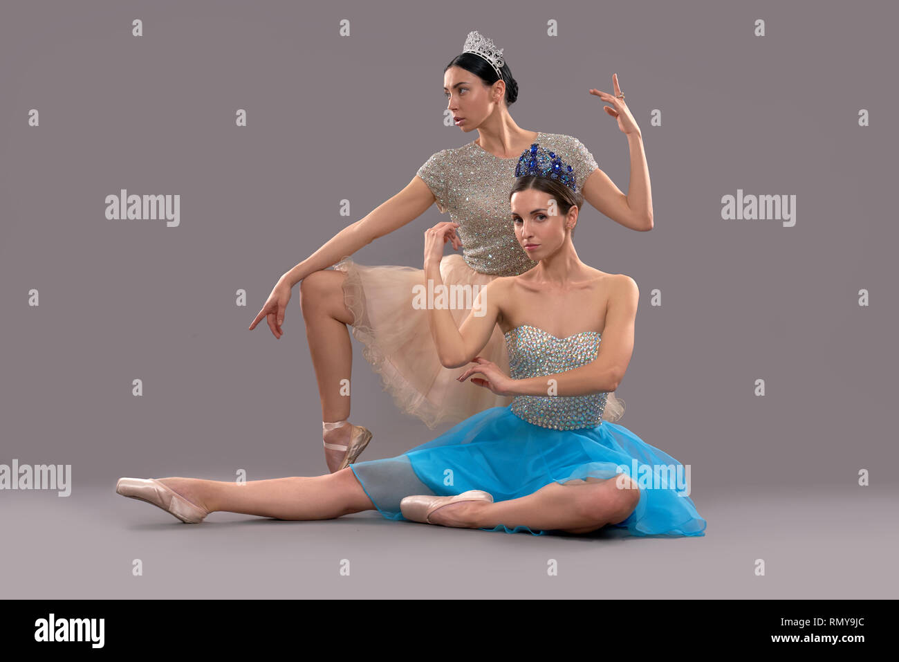 Due ballerine professioniste in posa sul grigio Sfondo isolato. Leggiadra  danzatrice in vestito blu seduti sul pavimento e guardando la telecamera  mentre la signora in abito color beige in posa dietro. Concetto