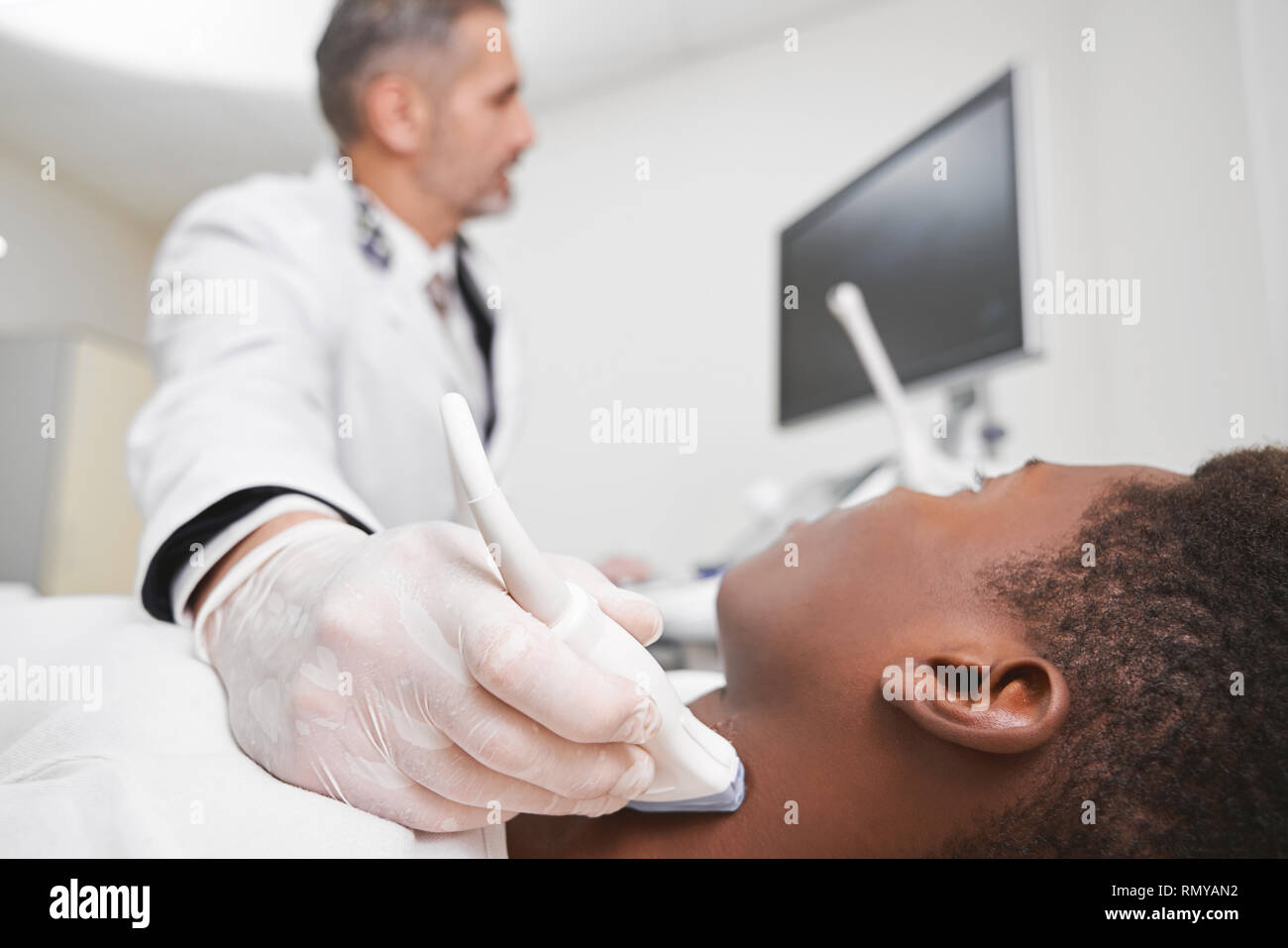 Medico tenendo la sonda ad ultrasuoni per diagnostisc ed esaminando i linfonodi sul collo dell'uomo. Paziente africano giacente nel gabinetto medico in clinica privata in montagna con attrezzature moderne. Foto Stock