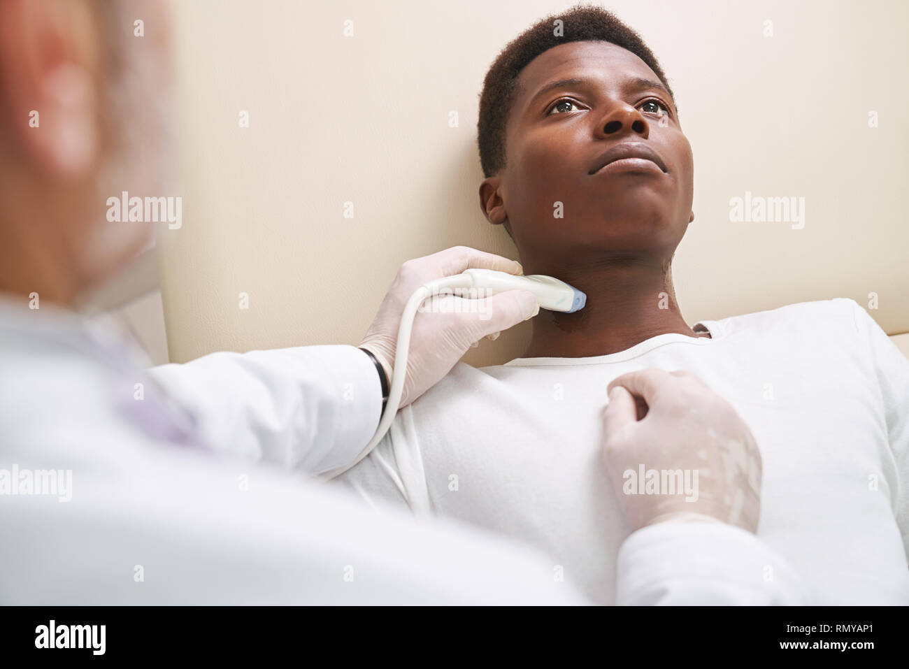 African paziente sdraiato, guardando verso l'alto. Medico di fare diagnosi ecografica, utilizzando, azienda speciale dispositivo. Sonda ad ultrasuoni per l'esame dei linfonodi sul collo. Terapista indossando guanti medicali. Foto Stock