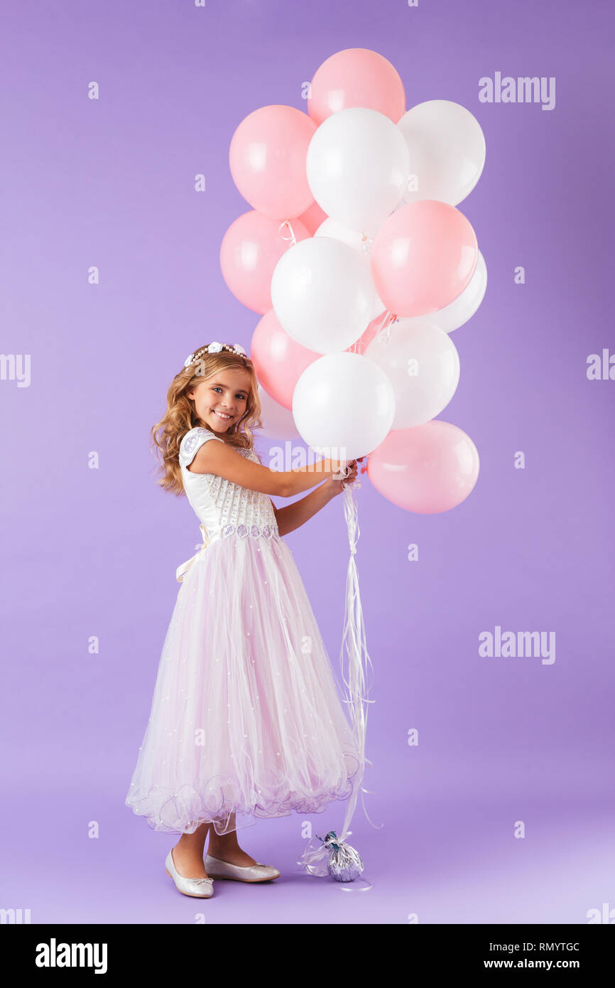 Lunghezza completa di una graziosa bambina vestito di principessa abito isolato in piedi su sfondo viola, tenendo mazzetto di palloncini Foto Stock